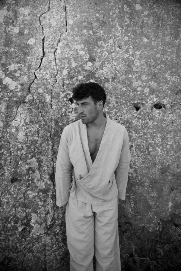 A man wearing a loose, light-colored kimono shirt made from natural fabric clothing stands against a weathered stone wall with visible cracks and small holes.